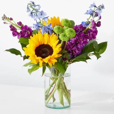 Bright sunflower, purple stock, blue delphinium, and green mums in a clear glass vase.