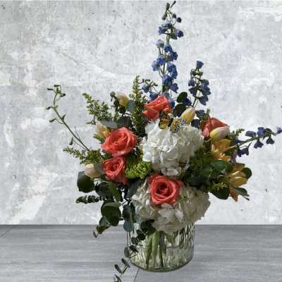 Mixed arrangement of white hydrangeas, coral roses, tulips, and blue delphinium in a clear glass vase
