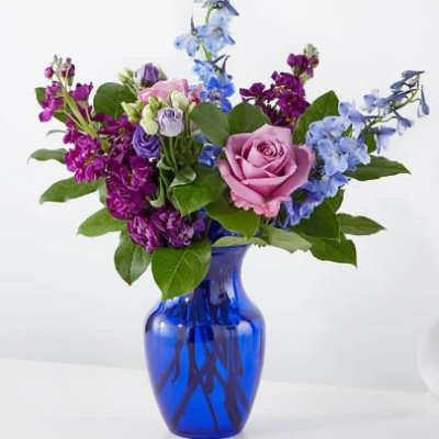 Purple, blue, and pink flower arrangement with roses and delphinium in a cobalt blue glass vase