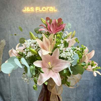 Pink lilies and baby's breath in a burgundy vase with a gold ribbon