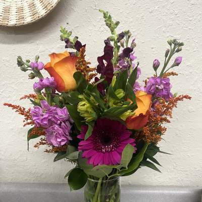 Mixed bouquet with orange roses, purple blooms, and a magenta gerbera in a glass vase