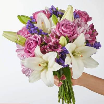 Hand-tied bouquet of white lilies, pink roses, and purple filler flowers held against a light background
