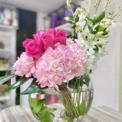 Round glass vase with pink roses, pink hydrangeas, and white orchids arranged low with visible stems and stones.