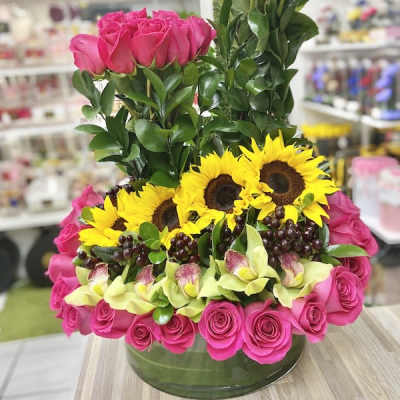 Structured arrangement of hot pink roses, yellow sunflowers, green orchids and grapes in a low glass vase