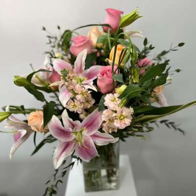 Tall arrangement of pink lilies, pink and peach roses, and pale stock in a clear glass vase.