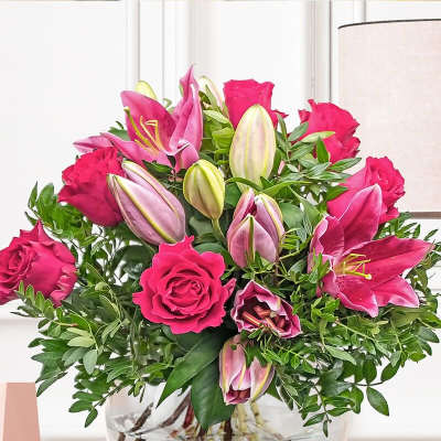 Arrangement of hot pink roses and pink lilies in a round clear glass vase on a wooden shelf
