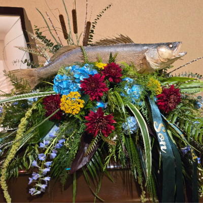 Fishing-themed casket spray with blue and red flowers, cascading ferns, and a mounted fish on top
