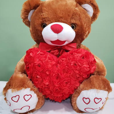 Brown teddy bear holding a red heart-shaped floral decoration