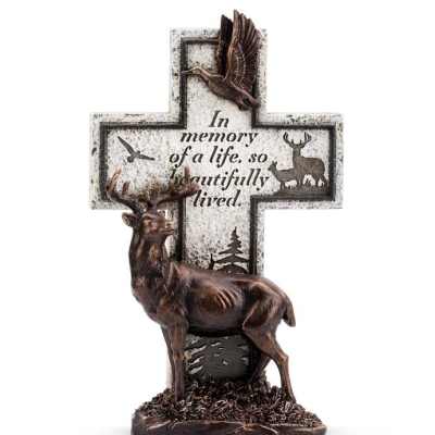Bronze-tone deer figurine in front of a stone-look memorial cross with bird and forest silhouettes and an inscription.
