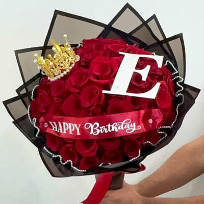 Bouquet of red roses wrapped in black paper with a crown, letter E, and Happy Birthday ribbon.