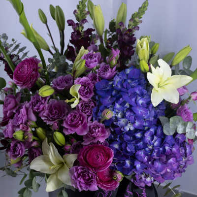 Purple and blue floral arrangement with white lilies in a black hat box