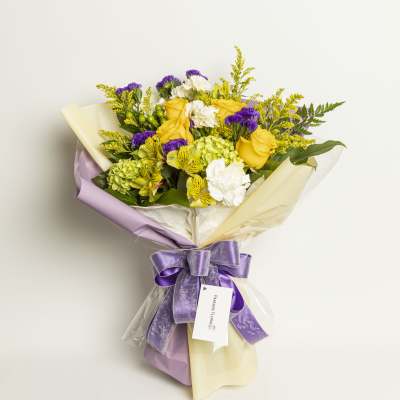 Bouquet of yellow roses, white flowers, and purple accents wrapped in lavender and cream paper