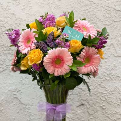 Pink gerbera daisies and yellow roses in a glass vase with a purple ribbon