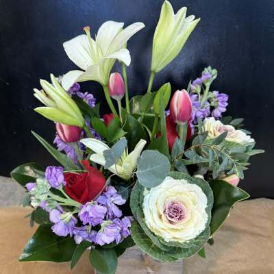 Mixed bouquet with lilies, roses, and purple blooms in a clear square vase