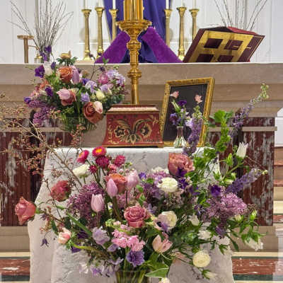 Large purple and pink mixed flower arrangements on a church altar with candles and framed photo