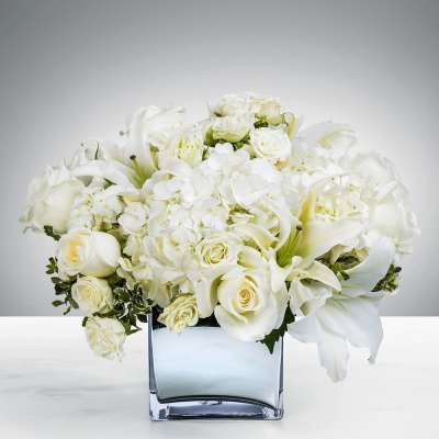 Compact arrangement of white roses, hydrangeas, and lilies in a square glass vase