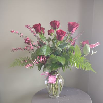 Tall arrangement of red roses with small pink flowers in a clear glass vase on a round table.