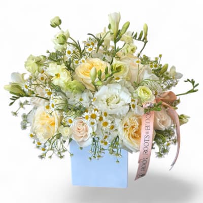 Low arrangement of white and pale yellow roses, daisies, and mixed blooms in a light blue cube vase with ribbon.