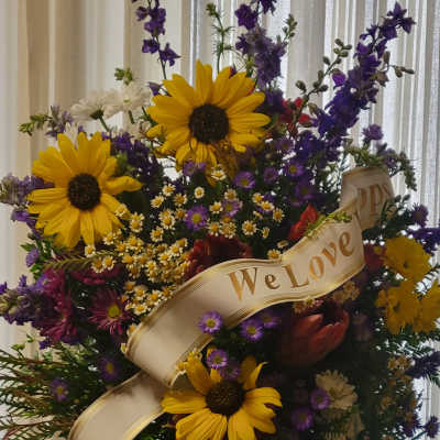 Tall floral standing spray with sunflowers, purple blooms, and a "We Love" ribbon