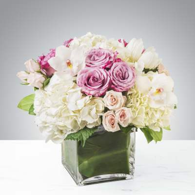 Low arrangement of pink roses, white hydrangeas, and white orchids in a square glass vase.