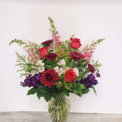 Tall mixed arrangement of red roses, burgundy mums, pink snapdragons and purple blooms in a clear glass vase