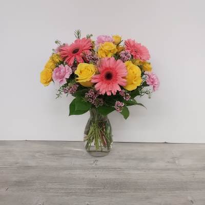 Pink gerbera daisies, yellow roses, and pink carnations arranged in a clear glass vase