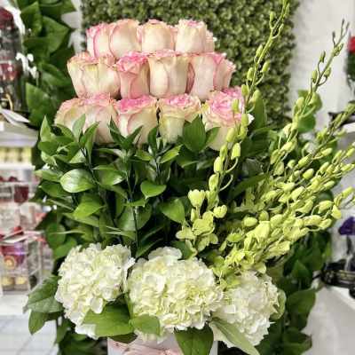Tall arrangement of pink-tipped roses, white hydrangeas, and green orchids in a white hatbox with pink ribbon