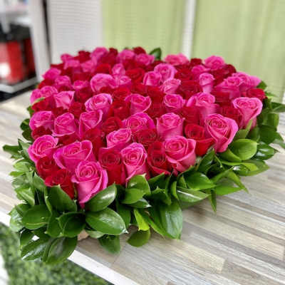 Heart-shaped arrangement of pink and red roses edged with glossy green foliage on a wooden surface