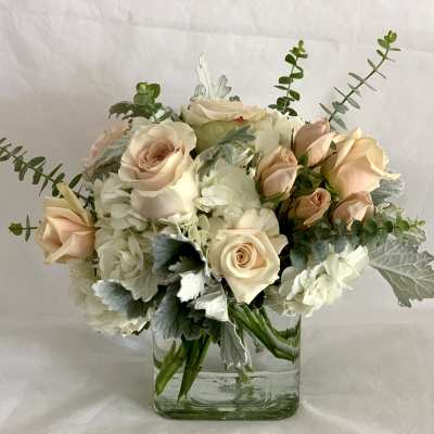 Low arrangement of cream hydrangeas and blush roses in a clear glass cube vase with soft gray foliage.