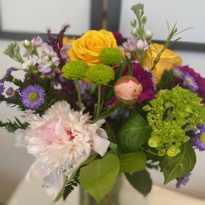 Mixed bouquet of yellow roses, pink peonies, and purple flowers in a glass vase