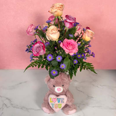 Pink and peach roses in a teddy bear vase with a plush bear holding a heart