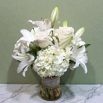 White lilies and hydrangeas arranged in a glass vase with lace trim