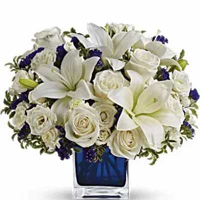 Low arrangement of white lilies and roses with purple accents in a blue cube glass vase