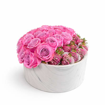Round white box filled with pink roses and chocolate-covered strawberries drizzled with pink and white.