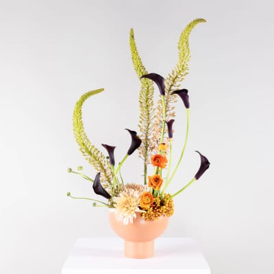 Tall floral arrangement with orange blooms and dark calla lilies in a peach vase