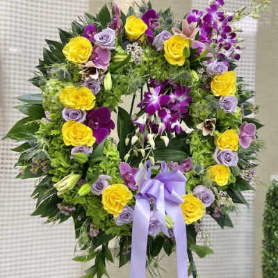 Standing wreath of yellow roses, lavender roses, and purple orchids with a lavender bow on an easel.