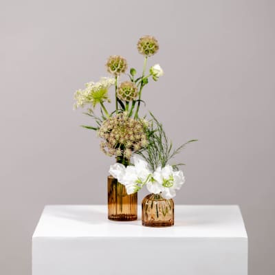 Two small floral arrangements in amber glass vases on a white pedestal
