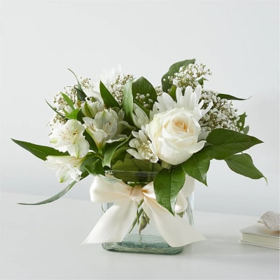 White roses and daisies arranged in a glass vase with a ribbon