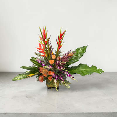 Tropical arrangement with orange roses, protea, purple orchids, and red heliconia in a low glass vase