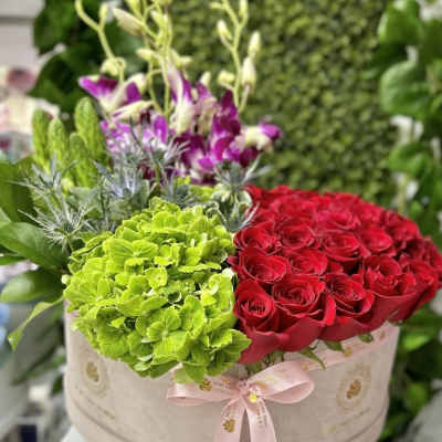 Round hatbox arrangement with red roses, green hydrangeas, and purple orchids tied with a pink ribbon.