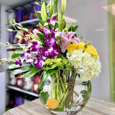 Tall mixed arrangement with white calla lilies, purple orchids, pink lilies, and yellow blooms in a round glass vase