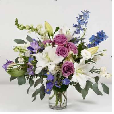 Arrangement of lavender roses, white lilies, and blue flowers in a clear glass vase