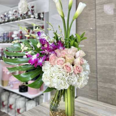 Tall mixed arrangement with white hydrangeas, pink roses, purple orchids and calla lilies in a clear glass vase
