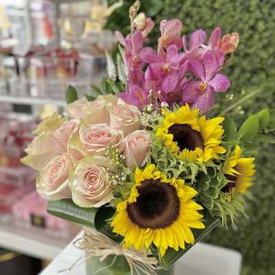 Arrangement of pale pink roses, yellow sunflowers, and pink orchids in a glass vase tied with raffia.