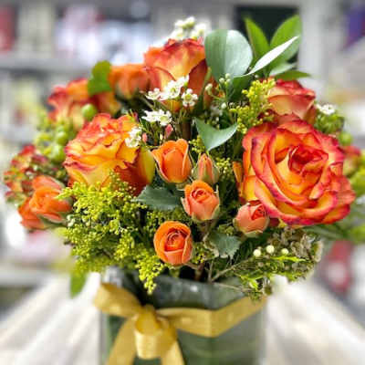 Orange and yellow roses with small white flowers in a leaf-lined square glass vase with a gold ribbon bow.