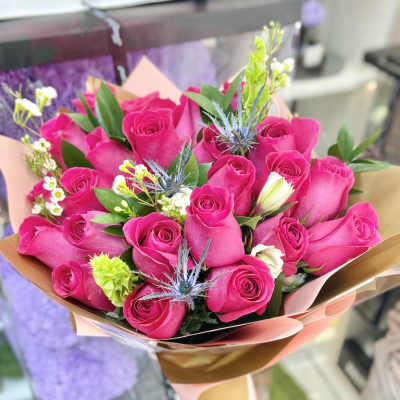 Hand-tied bouquet of bright pink roses with small white blooms and blue thistles wrapped in pink and gold paper