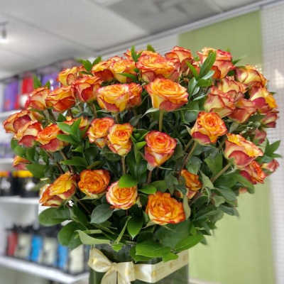 Large arrangement of orange and yellow roses in a square glass vase with a cream ribbon bow.