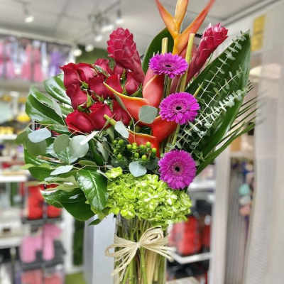 Tall tropical arrangement with red roses, orange blooms, green hydrangea and purple daisies in a glass vase