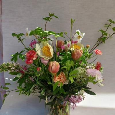 Tall garden-style arrangement of pink, peach, and white flowers with branches in a clear glass vase.