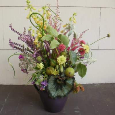 Wild-style arrangement of yellow and purple flowers with green fruits in a dark purple vase